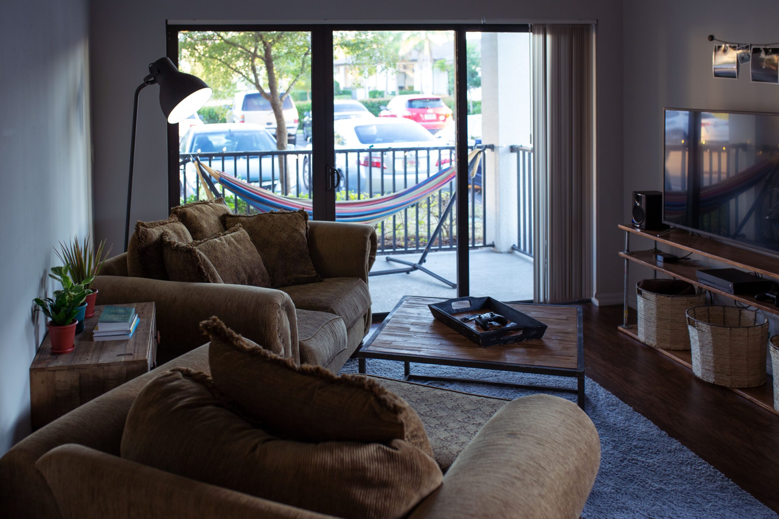 A warm and cozy apartment living room featuring comfortable brown sofas, a wooden coffee table, a floor lamp, and a large sliding glass door showing a balcony with a hammock and outdoor view.